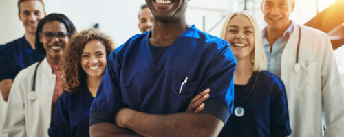 Young African male doctor smiling while standing in a hospital corridor with a diverse group of staff in the background